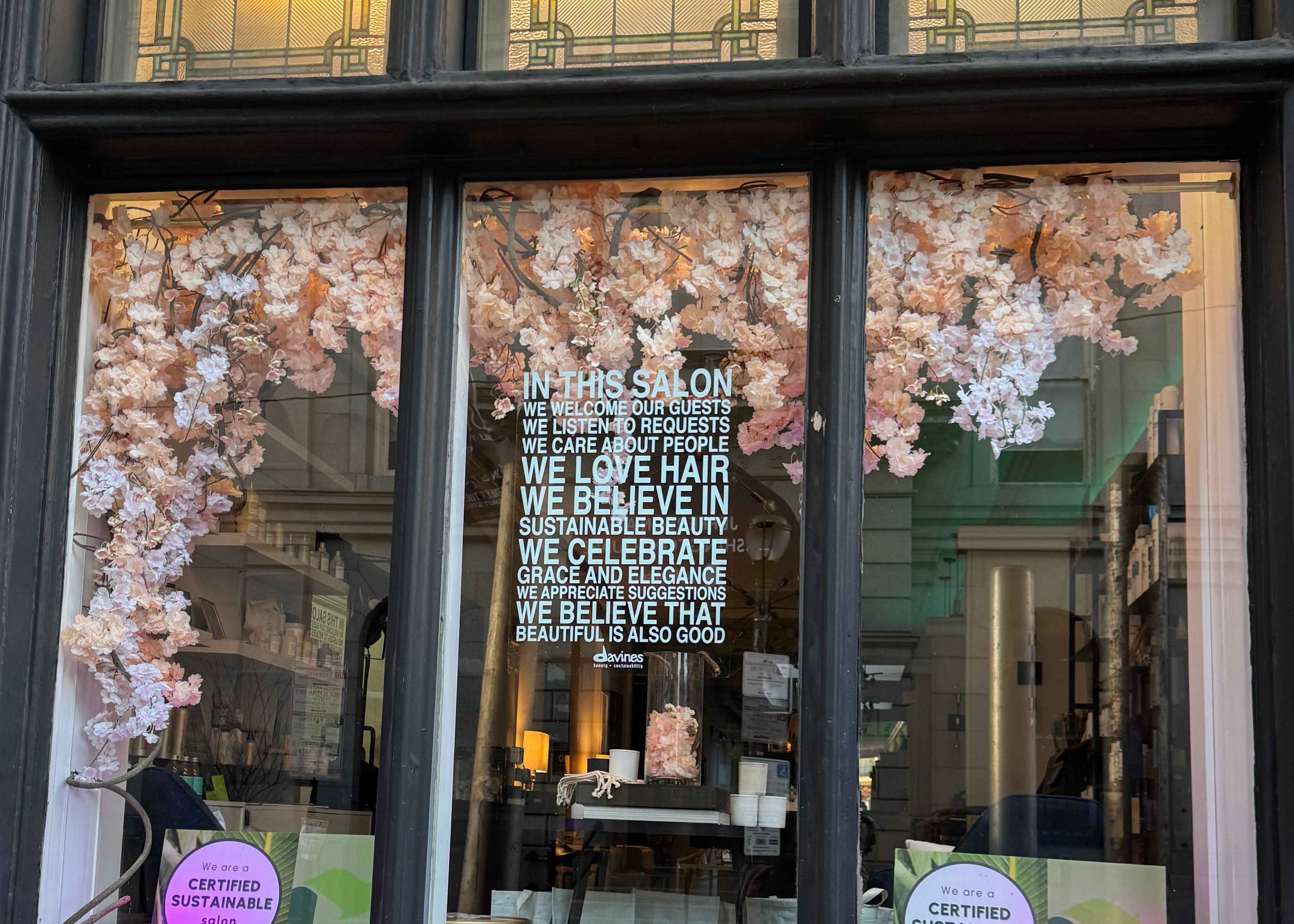 Window display with floral decorations and a sign inside a salon.